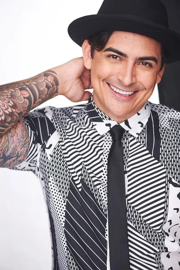 Portrait of Robbie Daymond smiling, wearing a black and white patterned shirt and a black hat.