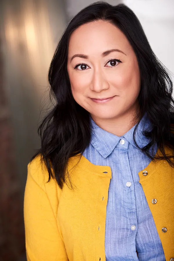 Portrait of Anne Yatco smiling, wearing a yellow cardigan over a blue shirt.