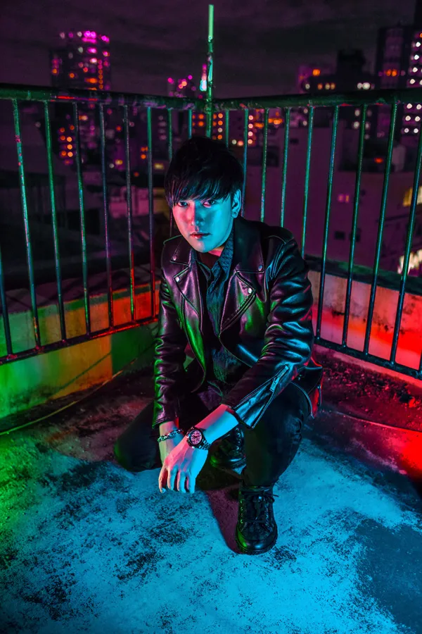 Portrait of TeddyLoid in a leather jacket against a neon-lit city background.