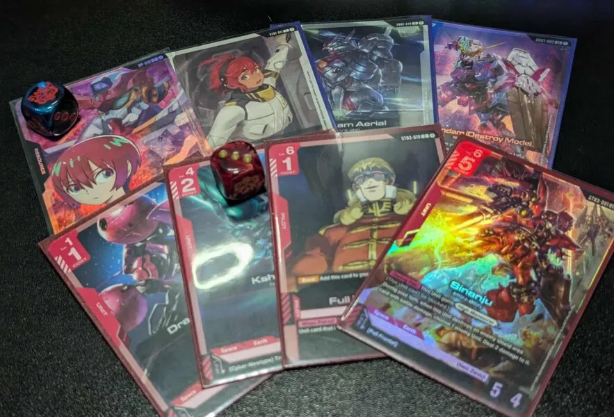 A collection of Gundam-themed trading cards and colorful dice spread out on a dark surface.