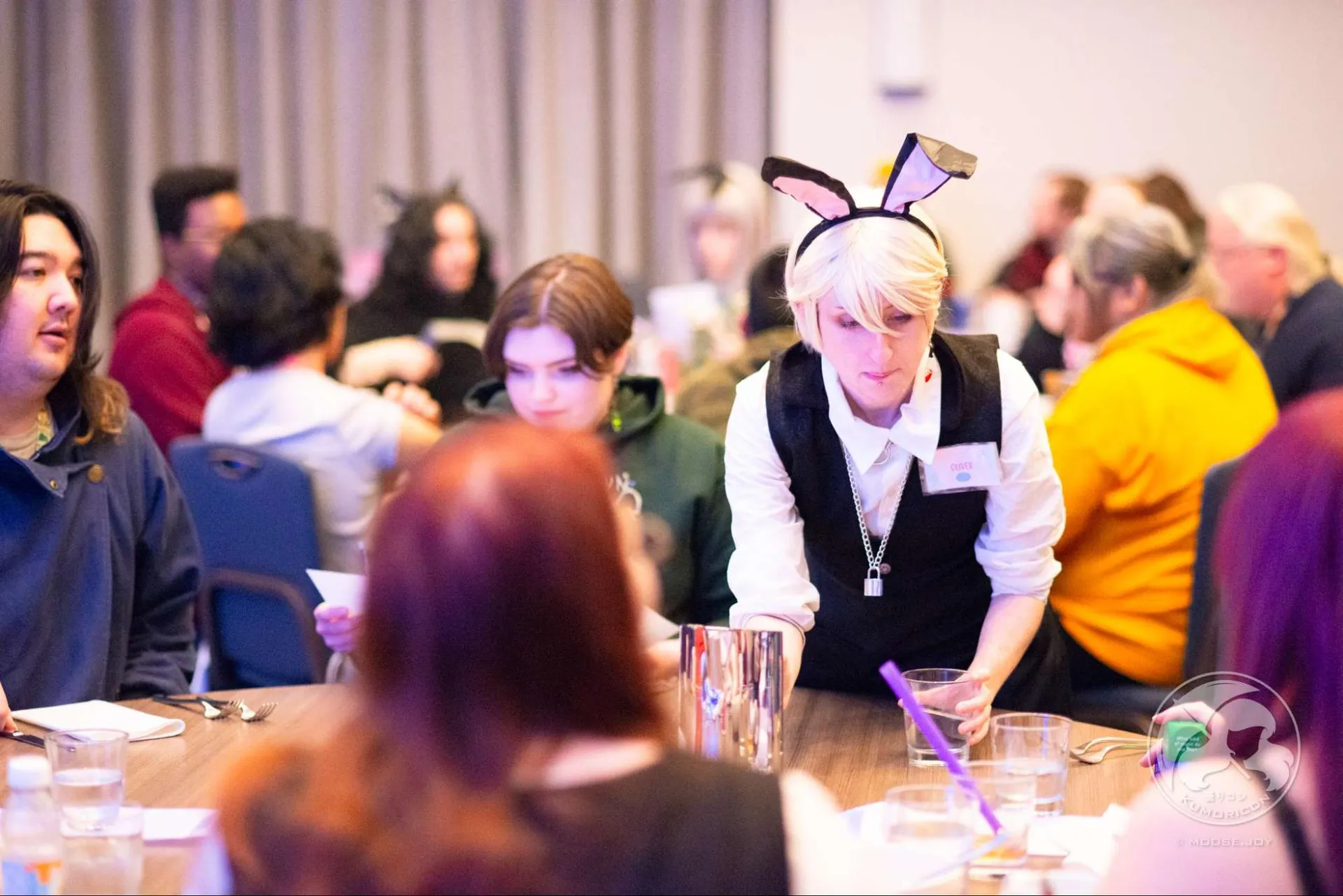 A maid cafe server in a black vest and bunny ears serving drinks to guests at a table.