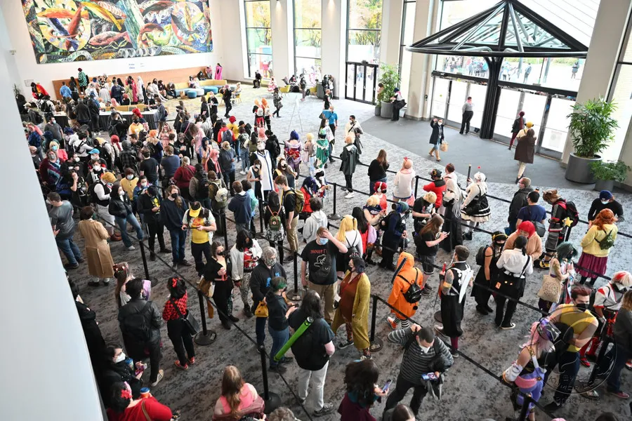 A high-angle view of a large, crowded convention hall with attendees in various costumes lined up for a health check.