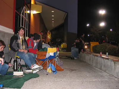 A photo of people waiting in line outside a store at night, sitting in camp chairs.