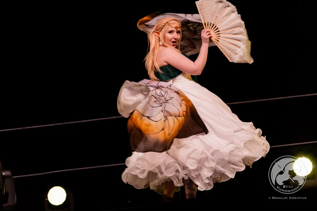 A performer in a Zelda cosplay gracefully dancing with a fan on stage.